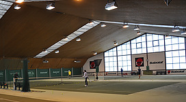 Tennishalle Braunau am Inn in Braunau am Inn: Es handelt sich um eine große, helle Sporthalle mit einem hohen Dach und zahlreichen Scheinwerfern. Der Boden besteht aus Hartplatz, auf dem sich einige Personen befinden, die vermutlich Tennis spielen oder trainieren. An den Wänden sind Werbeplakate zu sehen, die einen Sportartikelhersteller bewerben.