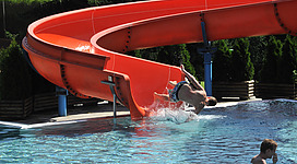 Ein Schwimmbad mit einer großen, orangefarbenen Rutsche. Ein Junge springt ins Wasser, während ein anderer im Pool schwimmt.