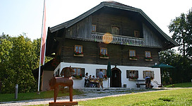 Ein traditionelles Holzhaus mit einem Garten in einer grünen Umgebung. Vor dem Haus steht eine Skulptur eines Hundes und es sind Gäste im Freien.