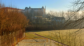 Beach-Volleyballplatz in Grein: Ein historisches Gebäude mit traditioneller Architektur erhebt sich über einer geräumigen, ländlichen Umgebung. Ein Kiesweg führt auf das Gebäude zu, umgeben von blühenden Büschen und nackten Bäumen. Die Landschaft ist von sanften Hügeln und einem klaren Himmel geprägt.