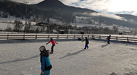 Ein Eislaufplatz in einer malerischen Winterlandschaft. Kinder fahren fröhlich auf dem Eis, umgeben von schneebedeckten Bergen.