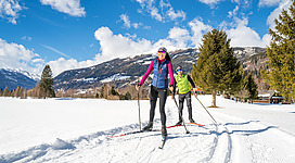 Zwei Personen fahren auf Langlaufskiern durch eine schneebedeckte Landschaft. Im Hintergrund sind Berge und ein klarer blauer Himmel zu sehen.