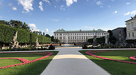 Ein eleganter Garten mit gepflegten Beeten und einem beeindruckenden Gebäude im Hintergrund. Der Himmel ist klar und einige Wolken sind sichtbar.