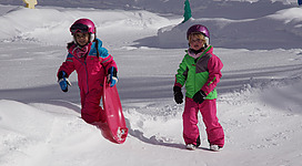 Zwei Kinder im bunten Schneeanzug spielen im Schnee. Sie haben Schlitten in der Hand und genießen einen sonnigen Wintertag.