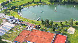 Eine Luftaufnahme zeigt einen Tennisplatz und einen Teich mit Freizeitmöglichkeiten. Umgeben von grünen Wiesen und Bäumen wirkt die Landschaft einladend und freundlich.