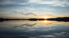 Europareservat Unterer Inn in Mining: Eine idyllische Seenlandschaft bei Sonnenuntergang. Der See spiegelt den orangeroten Himmel und die dunklen, bewaldeten Ufer wider. Auf der einen Seite ist eine kleine Insel zu sehen. Die ruhige Oberfläche des Sees lässt eine friedliche und entspannende Stimmung entstehen.