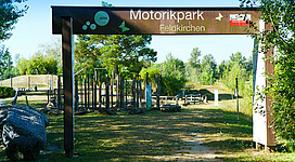 Motorikpark® in Feldkirchen an der Donau: Ein großer, offener Freizeitpark mit verschiedenen Spielgeräten und Einrichtungen. Der Haupteingang ist mit einem Schild gekennzeichnet, das den Namen "Motorikpark Feldkirchen" anzeigt. Die Umgebung ist von grünen Bäumen und Sträuchern umgeben, was eine angenehme Atmosphäre schafft.