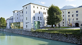 Ein modernes Gebäude neben einem Fluss, umgeben von Bäumen und Grünflächen. Die Architektur kombiniert historische und zeitgenössische Elemente.