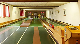 Kegelbahn - Rieder Wirt in Ried im Innkreis: Ein großer, heller Raum mit mehreren Bahnen für Bowlingspiele. Der Boden ist grün gestrichen, und an den Wänden hängen verschiedene Dekorationen. In der Mitte des Raums stehen mehrere Sitzgelegenheiten und am Ende der Bahn steht ein Scorepult.