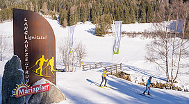 Ein Langlaufzentrum in einer verschneiten Landschaft mit einem großen Schild. Zwei Skifahrer fahren auf einer gespurten Strecke durch die Winterlandschaft.