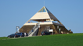 Pyramide am Etzinger Hügel in St. Agatha: Ein pyramidenförmiges Gebäude mit einem Spitzdach und hölzernen Konstruktionen. Es steht auf einer Erhebung inmitten einer grünen Wiese, wobei einige Autos in der Nähe geparkt sind. Die Architektur wirkt rustikal und traditionell.