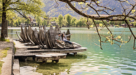 Gschmå Platzl Seerosen in Strobl: Das Bild zeigt einen idyllischen See, an dessen Ufer sich ein hölzerner Steg mit mehreren traditionellen Booten befindet. Im Hintergrund sind Bäume zu sehen, deren Zweige sich über den See erstrecken. Zwei Personen sitzen auf einer Bank auf dem Steg und scheinen die friedliche Umgebung zu genießen.