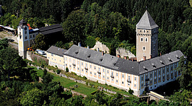 Schloss Neuhaus in St. Martin im Mühlkreis: Ein mehrstöckiges Gebäude mit einem hohen Turm, das von Bäumen umgeben ist. Die Fassade des Gebäudes weist eine klassische Architektur mit Dachgiebeln und Fenstern auf. Die Umgebung ist von dichter Vegetation und Berghängen geprägt, was eine malerische Szenerie ergibt.
