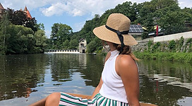 Zillenfahrt auf der Aist - "Hafen" in Schwertberg: Eine Person sitzt in einem Ruderboot auf einem ruhigen Fluss, umgeben von Bäumen und Vegetation. Sie trägt einen Strohhut und ein gestreiftes Oberteil, und schaut auf die Landschaft vor ihr. Das Boot wird von der Strömung des Wassers angetrieben.