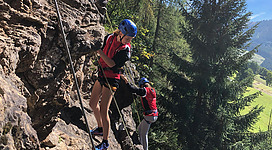 Zwei kletternde Personen an einer Felswand umgeben von Bäumen. Die Sonne scheint, und die Landschaft ist grün und bergig.
