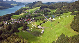 Eine malerische Landschaft mit sanften Hügeln, grünen Wiesen und einem glitzernden See. Im Hintergrund sind kleine Häuser und Wälder zu sehen.