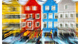 Alfred-Kubin-Galerie in Wernstein am Inn: Eine verschwommene Sicht auf ein Stadtbild. Mehrere farbenfrohe, traditionelle Gebäude sind zu erkennen, die in Bewegung erscheinen. Der Fokus liegt auf dem Kontrast zwischen den roten, blauen und grauen Fassaden. Es entsteht der Eindruck einer lebendigen, dynamischen Szene.