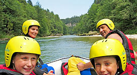 Eine Gruppe von vier Personen sitzt in einem Boot auf einem ruhigen Fluss. Alle tragen Helme und Schwimmwesten, um sicher zu bleiben.