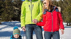 Eine Familie genießt einen Wintertag im Schnee. Der Vater und die Mutter halten die Hände, während ihr Kind auf einem Schlitten sitzt.