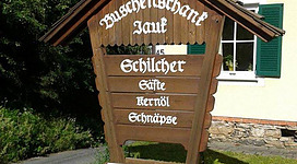 Ein rustikales Holzschild mit der Beschriftung "Buschenschank Jauß" und Angeboten wie Saft, Kernöl und Schnäpse. Es zeigt die Öffnungszeiten von Donnerstag bis Sonntag ab 14 Uhr.