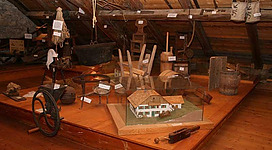 Ein Dachboden mit verschiedenen traditionellen Werkzeugen und Haushaltsgegenständen. In der Mitte steht ein Modell eines Hauses.