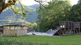 Skaterpark Kaltenbachau in Bad Ischl: Das Bild zeigt eine Parkanlage mit einer Betonfläche, die für Sport- und Freizeitaktivitäten genutzt wird. Darüber hindern ein hölzerner Unterstand und eine Rampe die Besucher zum Erkunden der Umgebung ein, die von Bäumen und Vegetation geprägt ist.