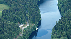 Ranna Stausee in Neustift im Mühlkreis: Eine weitläufige Berglandschaft mit einem breiten, stillen Fluss oder See, der sich durch dicht bewaldete Hänge schlängelt. Entlang der Ufer sind einige kleine Häuser und eine Straße zu sehen. Auf einer Anhöhe befindet sich eine Staumauer oder ein Damm. Die Umgebung wirkt malerisch und ruhig.