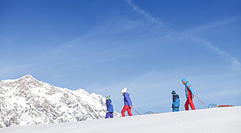 Eine Gruppe von Personen zieht Schlitten über einen schneebedeckten Hang. Im Hintergrund sind hohe Berge und ein klarer blauer Himmel zu sehen.