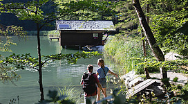 Ein ruhiger See umgeben von Bäumen und einer Hütte. Zwei Personen stehen am Ufer und genießen die Natur.