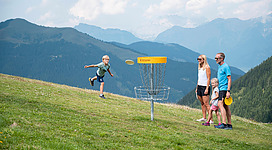 Eine Familie spielt Discgolf auf einer Bergwiese. Ein Junge wirft einen Frisbee in Richtung des Korbs, während die anderen zuschauen.