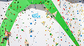 Eine Kletterwand mit vielen bunten Griffen und einer grünen Struktur. Ein Kletterer befindet sich in der vertikalen Bewegung.