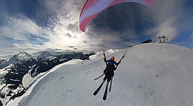 Ein Skifahrer mit einem Gleitschirm schwebt über einer schneebedeckten Berglandschaft. Der Himmel ist klar mit einigen Wolken.