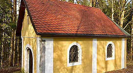 Grünbaumkapelle in Mauthausen: Eine kleine, gelbgetönte Kapelle mit rötlichem Ziegeldach steht inmitten eines bewaldeten Gebiets. Sie besitzt mehrere Bogenfenster mit dekorativen Gittermustern. Der Eingang ist durch einen Vorbau geschützt. Die Kapelle fügt sich harmonisch in die natürliche Umgebung aus Bäumen und Sträuchern ein.