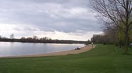 Hohenlohe AU-SEE in Enns: Eine grüne Rasenfläche erstreckt sich entlang eines Sees. Vereinzelte Bäume säumen den Uferbereich. Am Horizont sind leicht bewölkte Himmel zu erkennen.