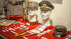 Ein Schaufenster mit historischen Militäruniformen und einer Auswahl von Mützen. Auf dem Tisch liegen verschiedene Orden und Medaillen.