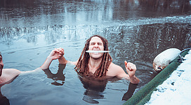 Eisbaden in Steyregg: Ein Mann schwimmt in einem schneebedeckten Teich und hält einen Stock in seinem Mund. Er hat lange Haare und scheint einen fröhlichen Gesichtsausdruck zu haben. Der Hintergrund zeigt eine winterliche Landschaft mit bewaldeten Ufern und einer ruhigen Wasseroberfläche.