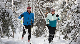Langlaufen in Kleinzell im Mühlkreis: Zwei Personen in winterlicher Kleidung, die auf Schneeschuhen durch einen schneebedeckten Wald laufen. Die Umgebung ist von hohen Bäumen umgeben, deren Äste mit Schnee bedeckt sind. Die Personen tragen warme Jacken und Handschuhe und scheinen die Winterlandschaft zu genießen.