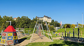 Generationen-Motorikpark® in Grein: Ein großer Spielplatz mit bunten Spielgeräten, Schaukeln und Klettergerüsten. Im Hintergrund ist ein malerisches, historisches Gebäude mit Türmen zu sehen, umgeben von grünen Bäumen. Kinder spielen fröhlich auf dem Spielplatz.