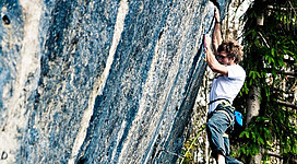 Ein Kletterer, der an einer steilen Felswand hochklettert. Die Umgebung ist grün und bewaldet.