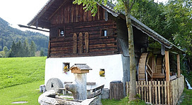 Ein traditionelles, rustikales Holzhaus mit einem weißen Untergeschoss und einem Wasserrad. Umgeben von grünen Wiesen und Bäumen.