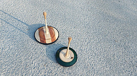 Zwei Holzkreisel stehen auf einem grauen Untergrund. Der linke Kreisel hat eine helle Holzoberfläche, während der rechte Kreisel eine grüne Basis hat.