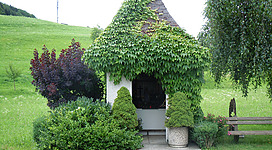 Binder Kapelle in Weyregg am Attersee: Das Bild zeigt ein kleines, gemütliches Gebäude mit einem Dach, das vollständig mit grünen Blättern bedeckt ist. Das Gebäude ist von üppiger Vegetation umgeben, darunter Bäume, Sträucher und blühende Pflanzen. Die Umgebung erscheint friedlich und idyllisch.