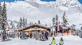 Ein belebtes Skigebiet mit einem charmanten Holzhaus und schneebedeckten Bergen im Hintergrund. Zwei Kinder in Ski-Ausrüstung laufen die Piste hinunter.