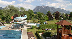 Ein Freibad mit großer Wasserrutsche, mehreren Becken und weitläufiger Liegewiese ist gut besucht. Menschen baden, rutschen oder entspannen im Schatten der Bäume. Im Hintergrund grüner Wald, Dächer und ein markanter Berg unter blauem Himmel mit Wolken.