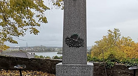 Kriegerdenkmal in Mauthausen: Ein hoher Obelisk aus Stein mit einem großen Eisenkreuz oben. Der Obelisk steht auf einem erhöhten Sockel und ist von Bäumen umgeben. Auf der Vorderseite des Sockels befinden sich Inschriften in einer anderen Sprache.