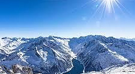 Eine beeindruckende Berglandschaft mit einem klaren See und grünen Wiesen. Die schneebedeckten Gipfel erheben sich majestätisch im Hintergrund.