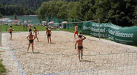 Ein Beachvolleyballfeld in der Natur mit mehreren Spielern. Im Hintergrund sind Bäume und ein See zu sehen.