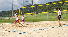 Drei Personen spielen Beachvolleyball auf einem Sandplatz. Im Hintergrund sind Bäume und ein bewölkter Himmel zu sehen.