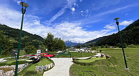 Ein schöner Park mit einem Weg, der zu einer Miniatureisenbahn führt. Im Hintergrund sind Berge unter einem klaren blauen Himmel zu sehen.