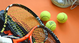 Ein Tennisracket liegt neben drei Tennisbällen auf einem orangefarbenen Untergrund. Im Hintergrund sind Sportschuhe zu sehen.